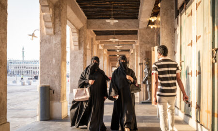 Două femei purtând hijab se plimbă pe unul dintre pasajele din souq, în timp ce un bărbat indian trece pe lângă ele și se uită la ele, Sursa foto dreamstime.com